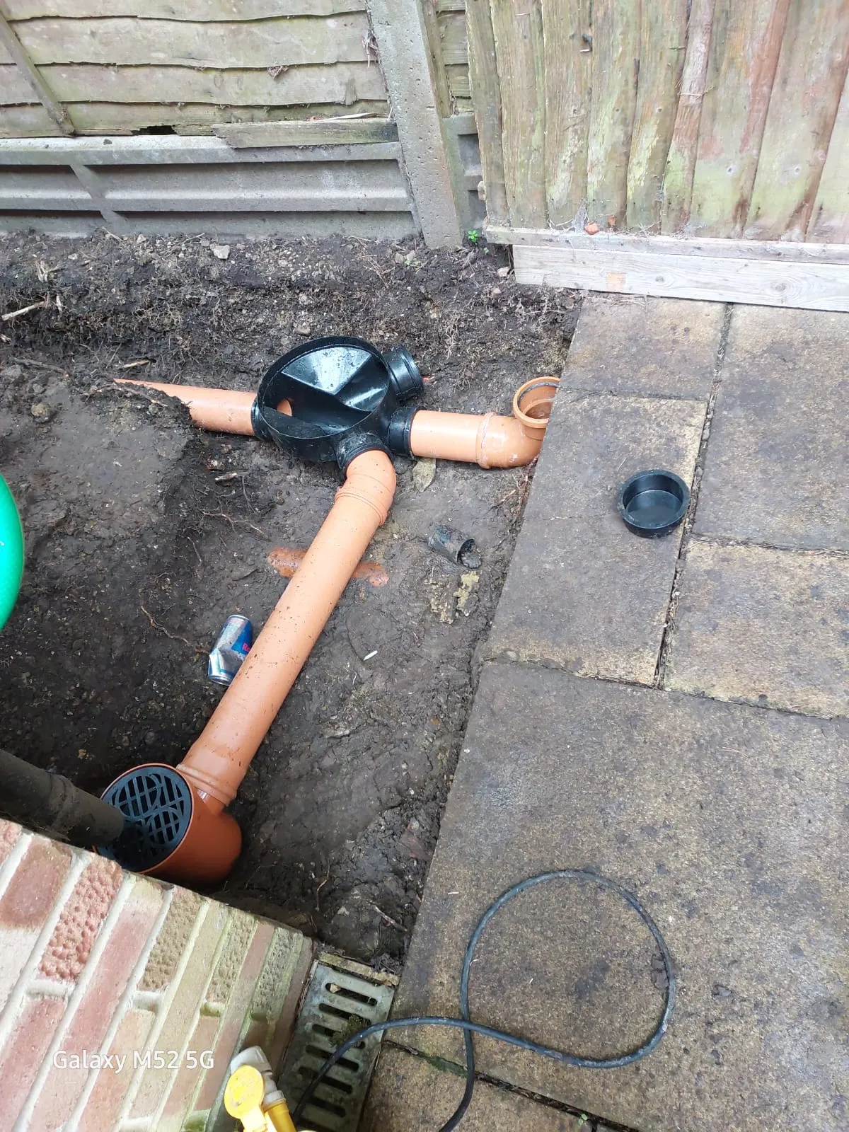 An outdoor trench showing new orange drainage pipes, a black inspection chamber, and a drain grate, indicating recent drain installation or repair work.