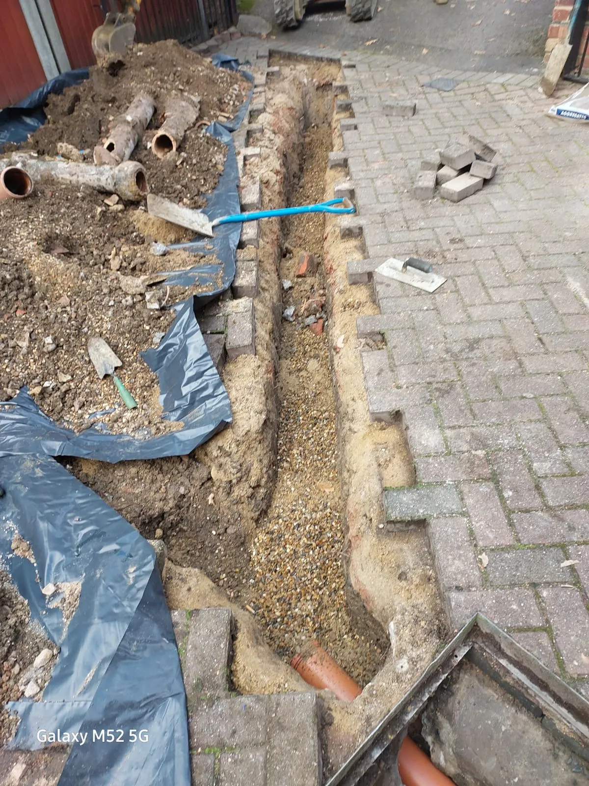 Excavated trench during drain repair and installation, with new orange pipes visible and old pipes removed, on a paved residential property.