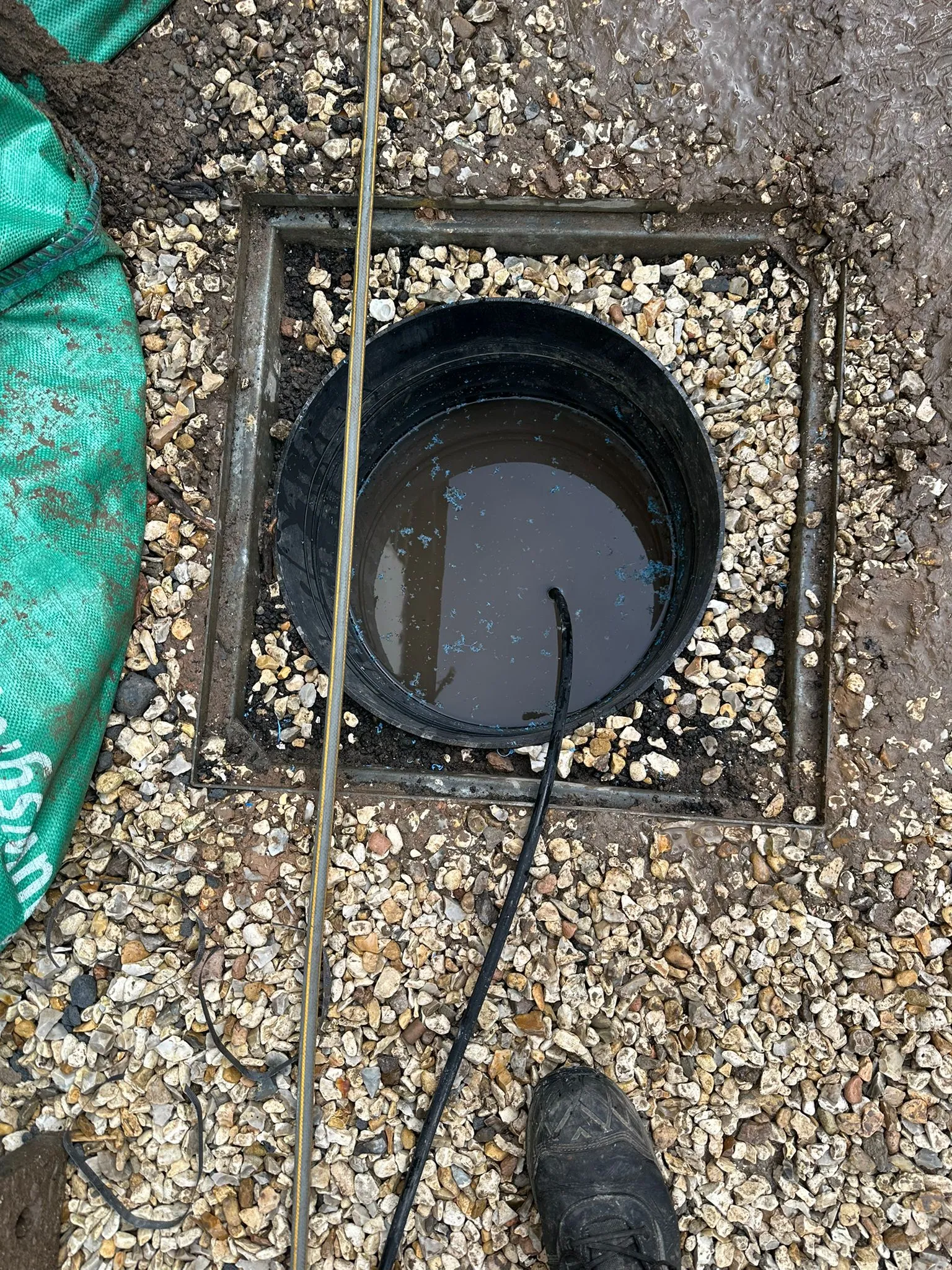 An outdoor drain access point with a severe blockage, showing murky water and a drainage hose or CCTV camera cable in use for emergency unblocking or inspection.
