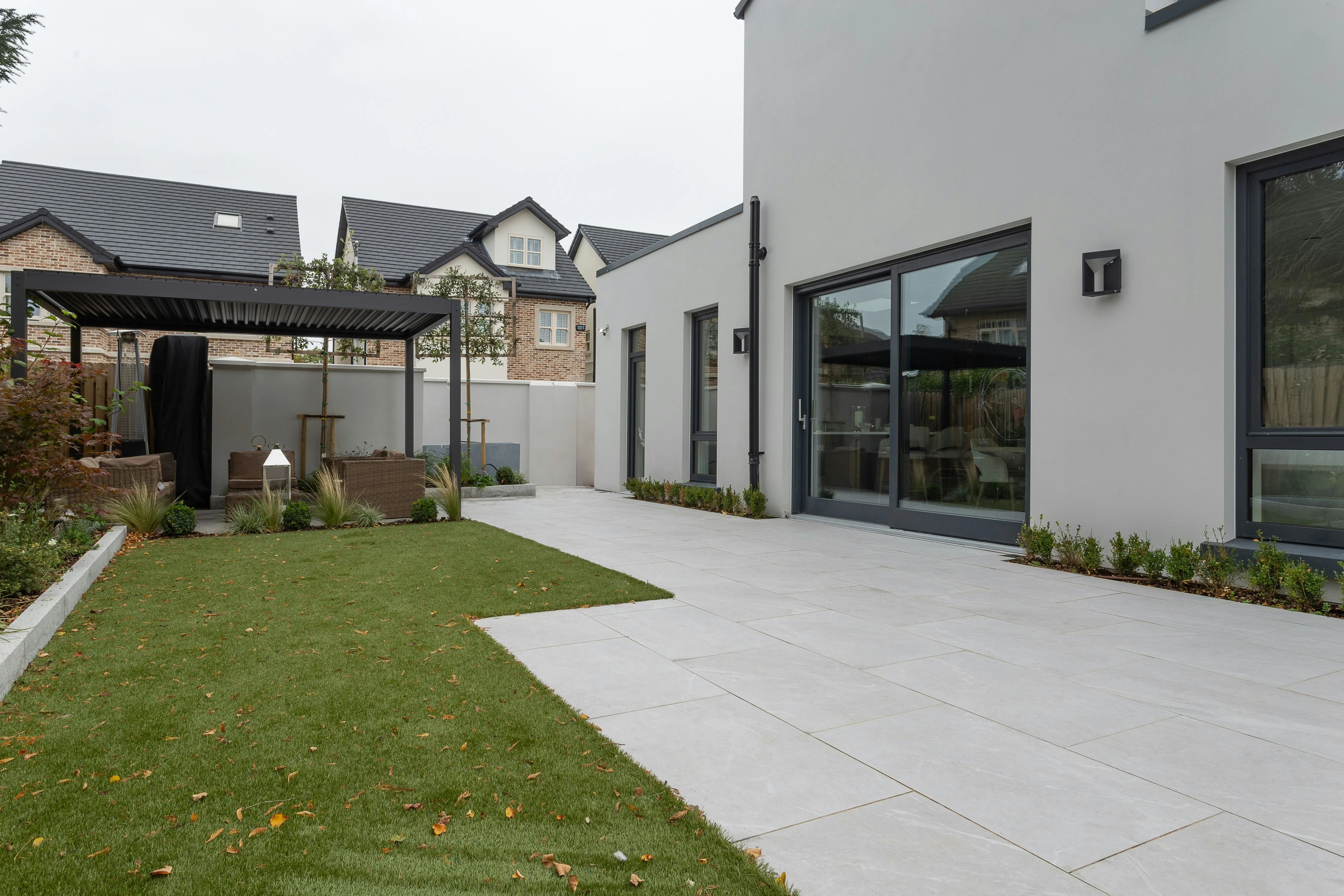 Modern garden patio with large format grey slabs, artificial lawn, and outdoor seating under a pergola.