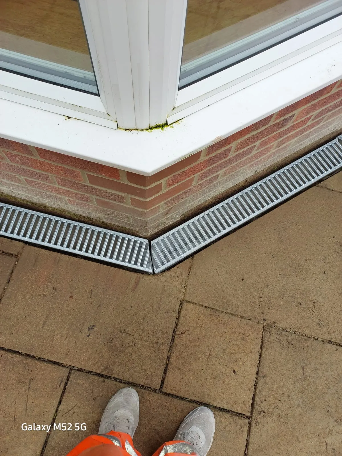 Installed ACO channel drainage system at the base of a building corner with a worker's feet visible.