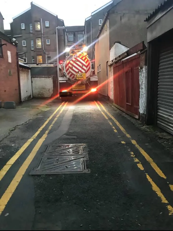 A drainage jetting and vacuum tanker truck working in a narrow alleyway or street, with a manhole cover in the foreground.