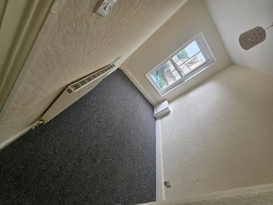 A clean, empty room after a house clearance, showing grey carpet, white textured walls, a white radiator, and a window looking outdoors.
