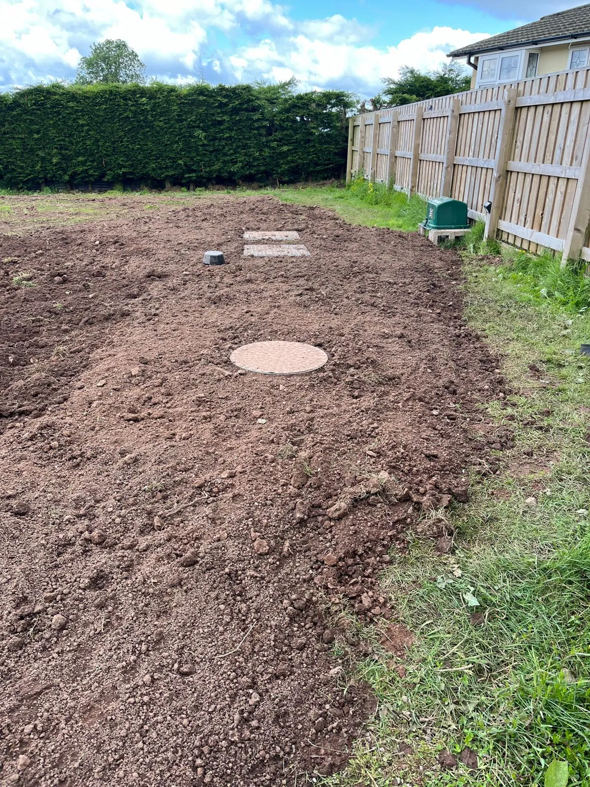 Garden groundwork with soil, circular manhole cover, other access points, and a green pump station unit near a fence.