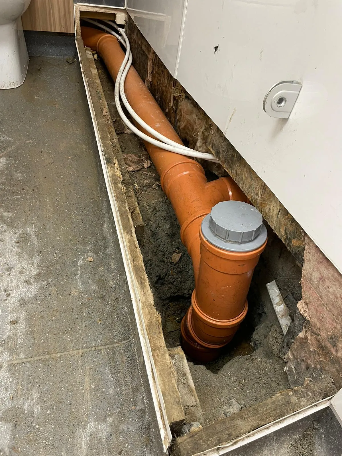 Orange plastic drainage pipes laid in an open trench beneath a floor, with a toilet visible on the left. The pipes are connected and appear to be new or recently repaired, with a grey cap visible on one section. White cables run alongside the pipes, and exposed earth and timber surround the installation.