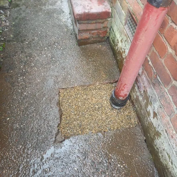 A picture of a blocked drain or gully with water overflowing onto the ground next to a residential brick wall and downspout.