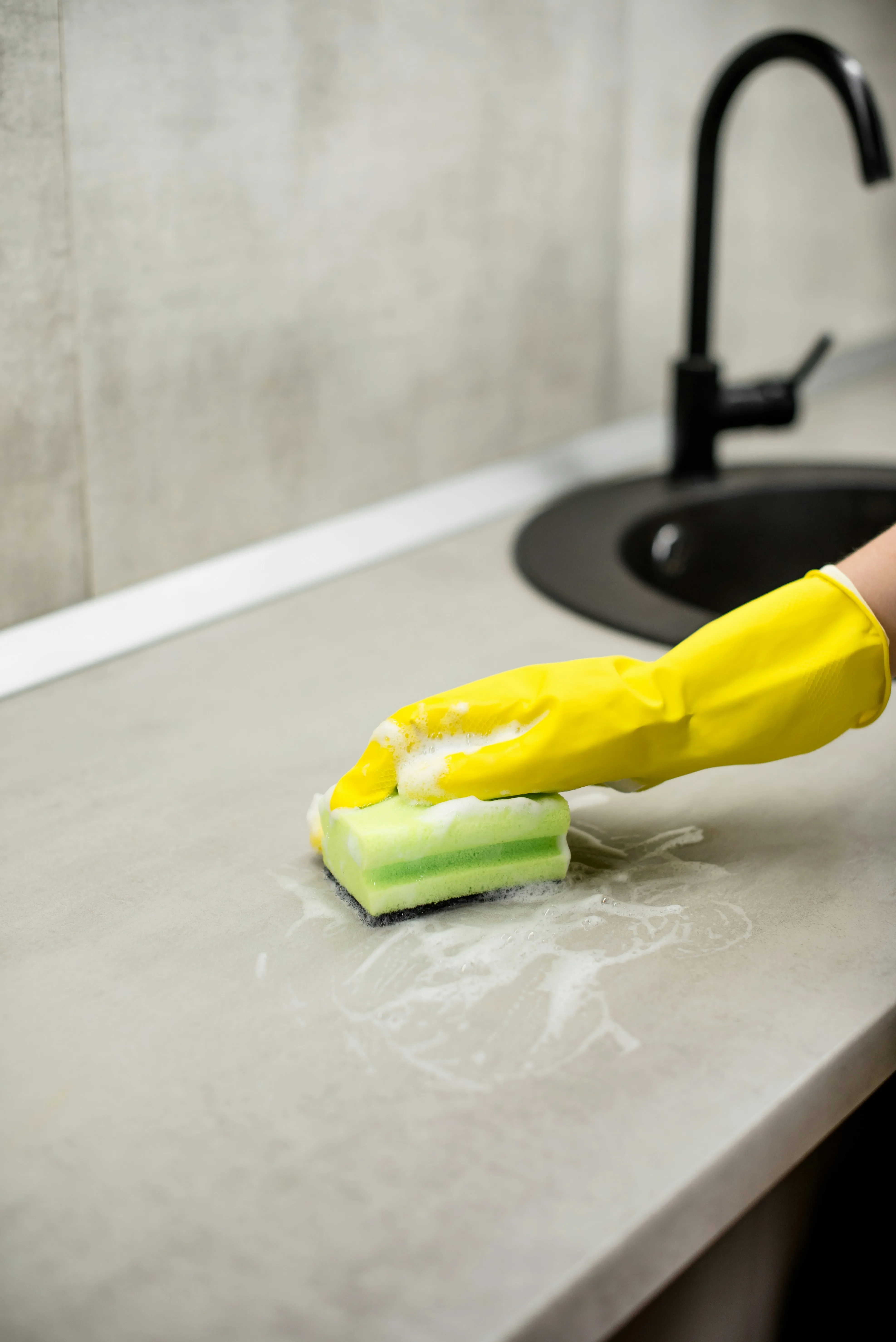 Person in yellow gloves scrubbing a modern kitchen counter with a green sponge