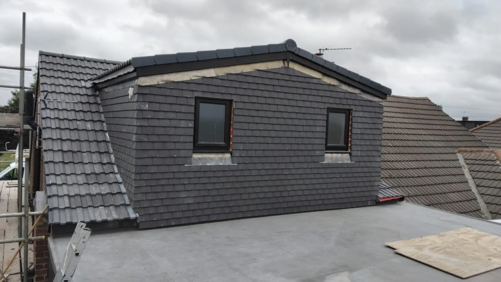 New dormer roof construction featuring dark grey tiles and windows, with a flat roof and scaffolding, illustrating comprehensive residential roofing services.