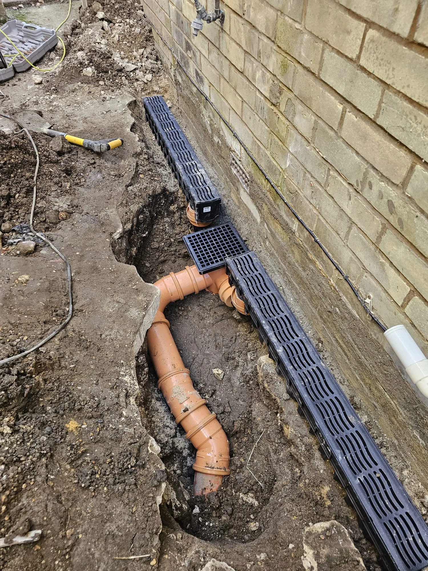 Trench next to a brick wall showing newly laid orange drainage pipes and black channel drains with grates, part of a new drainage system installation during groundworks.