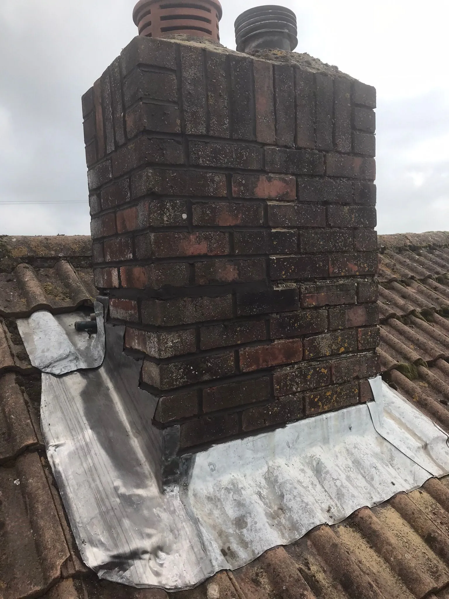 A close-up view of a brick chimney with lead flashing on a tiled roof, showing signs of wear and potential need for repair.