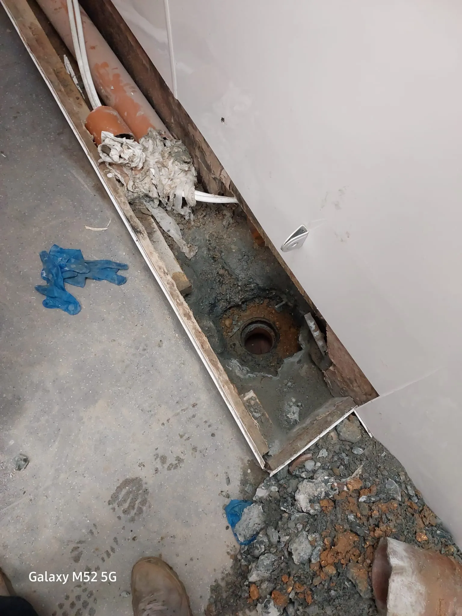 An open trench in a concrete floor revealing brown drainage pipes, a dark drain gully, and construction debris, suggesting drain repair or installation.