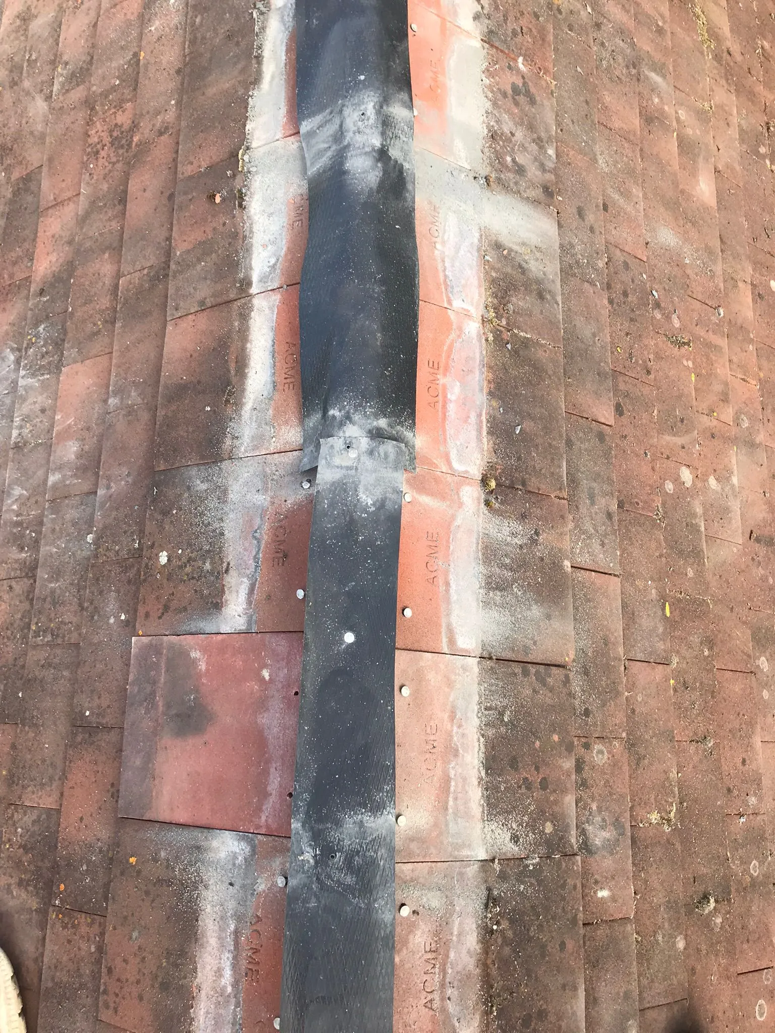 Weathered terracotta tiled roof valley with black flashing and visible fixings, showing roof maintenance.