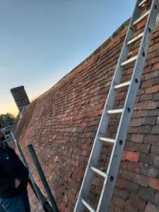A roofer on a ladder inspecting or repairing a terracotta-tiled pitched roof.
