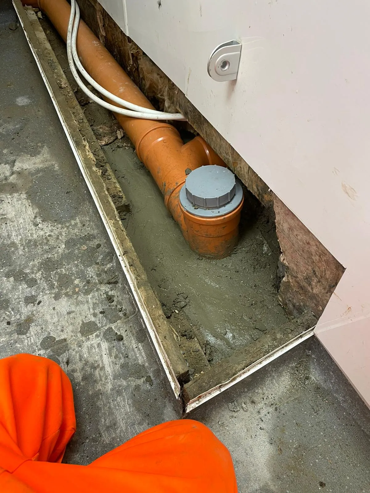Orange drainage pipes and electrical cables laid in a trench, partially covered with wet concrete during an installation or repair.