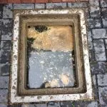 An outdoor drain with its cover removed, showing stagnant water and fat-like deposits indicating a blockage.