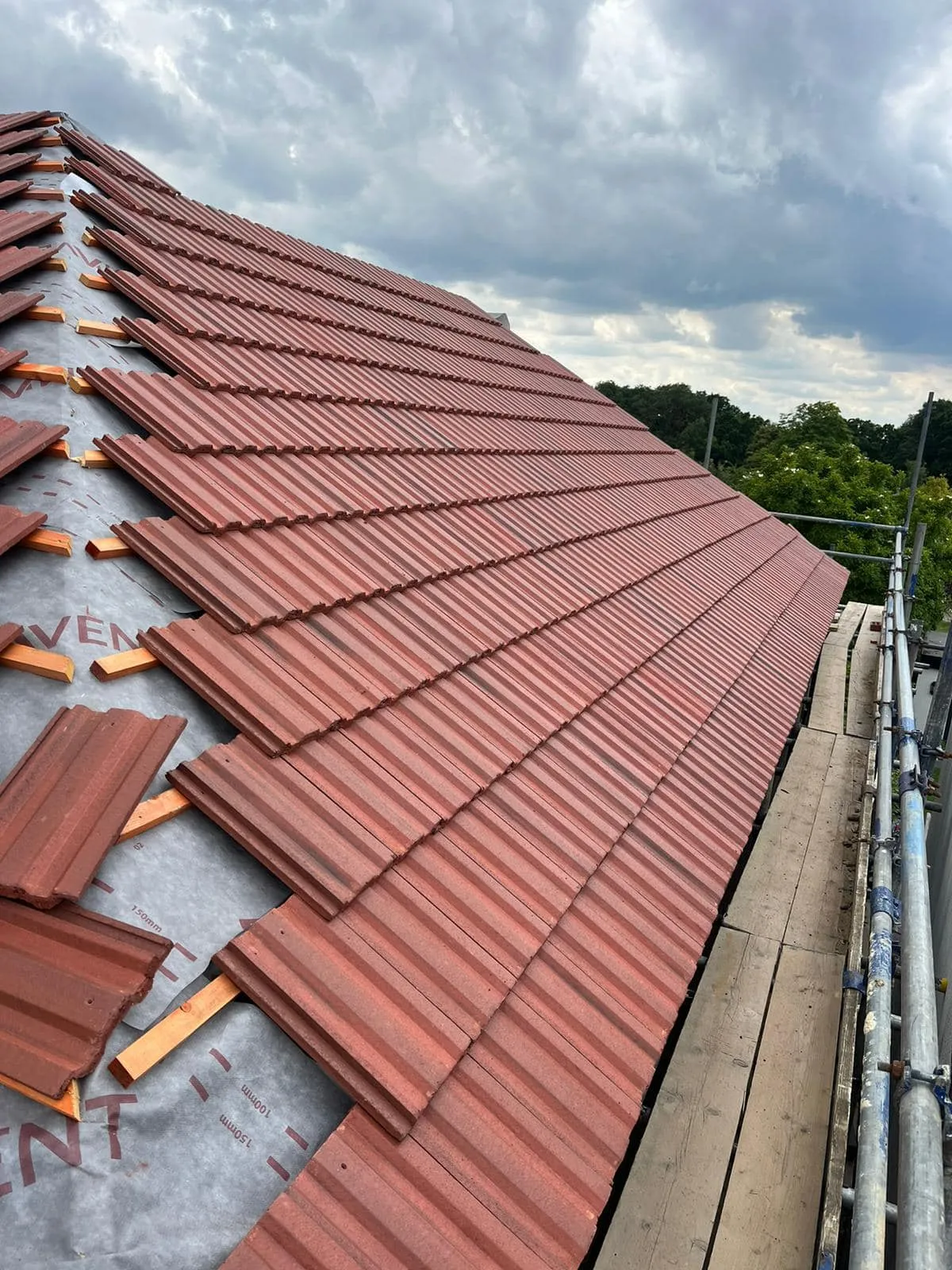 Professional roofers installing new concrete tiles on a pitched roof over a grey membrane, with scaffolding visible. Shows progress of a re-roofing project.