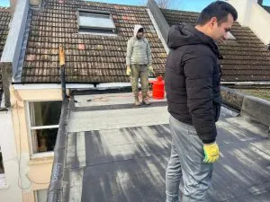 Two professional roofers on a flat roof, installing new dark roofing material. A tiled pitched roof with a skylight is behind them.