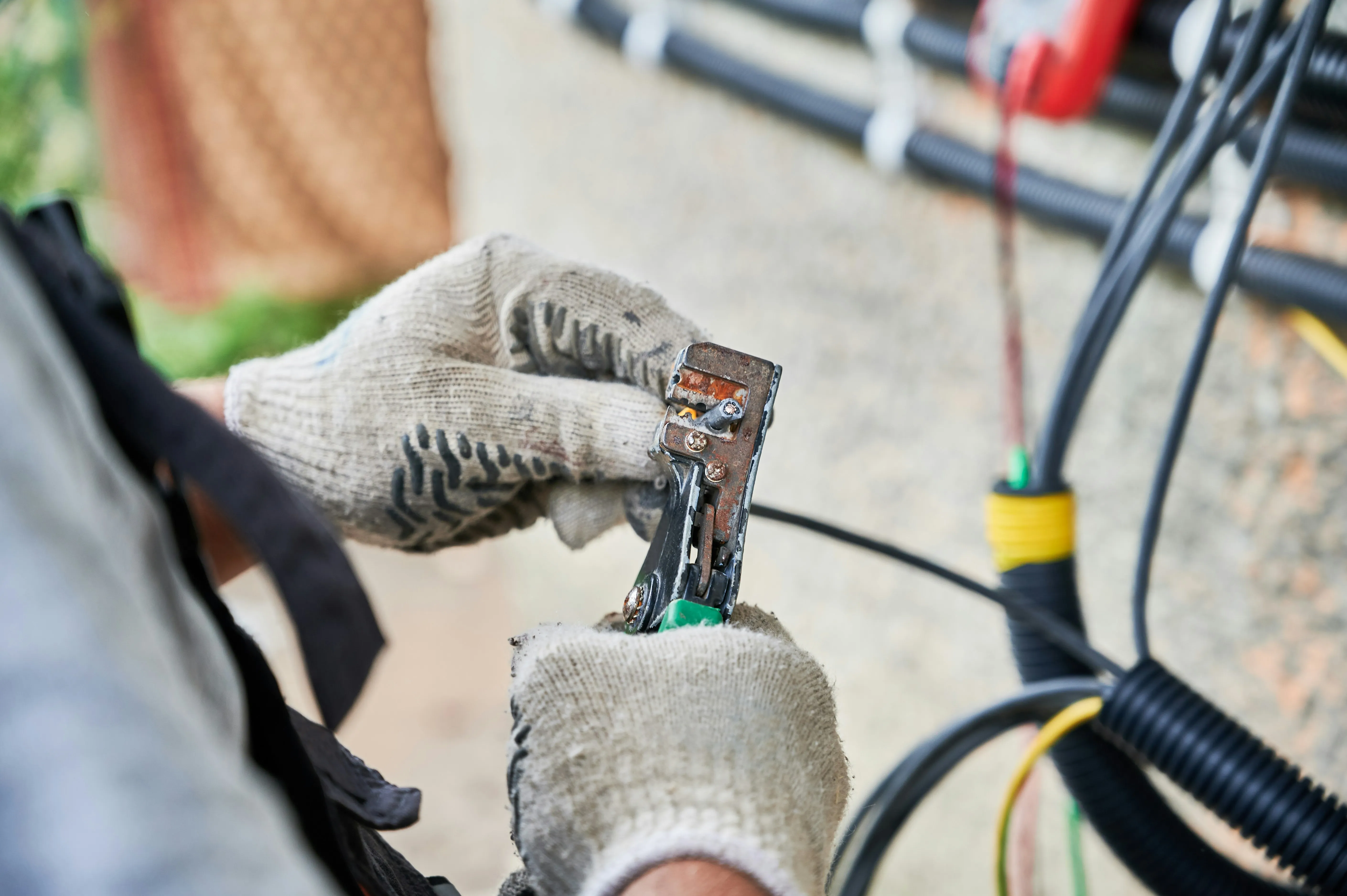 Electrician's gloved hands stripping an electrical cable with wire strippers.