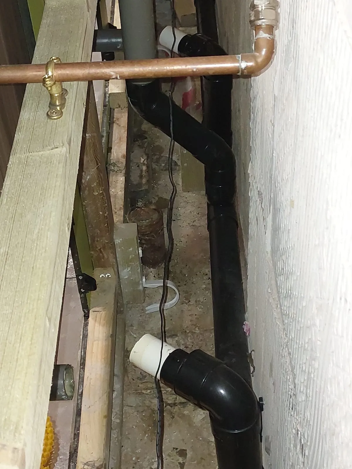 Newly installed black plastic drainage pipes and copper water pipes visible between wooden joists and against a wall during plumbing work.