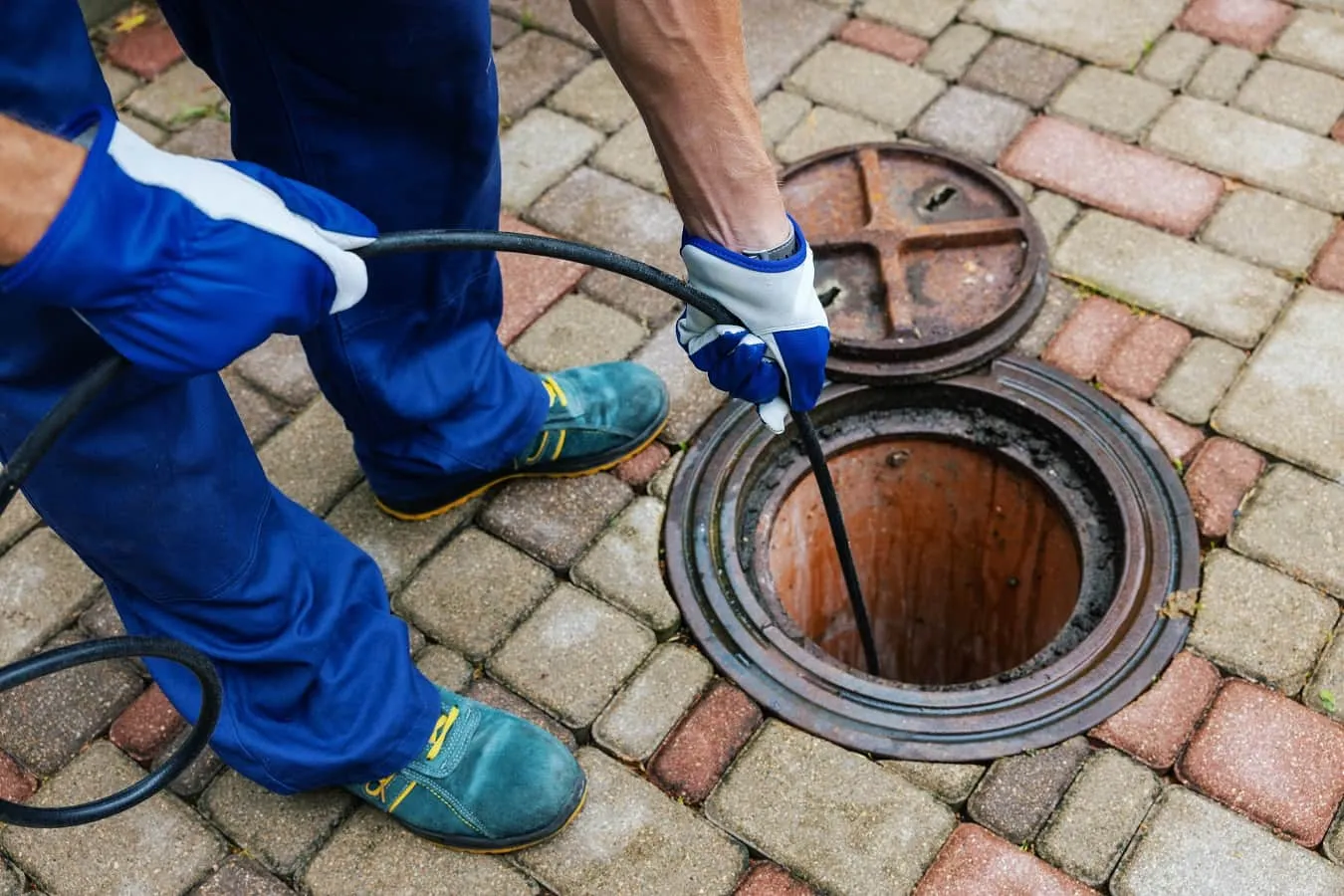 A person in blue uniform and gloves inserting a black flexible rod or cable into an open manhole cover on a paved surface.