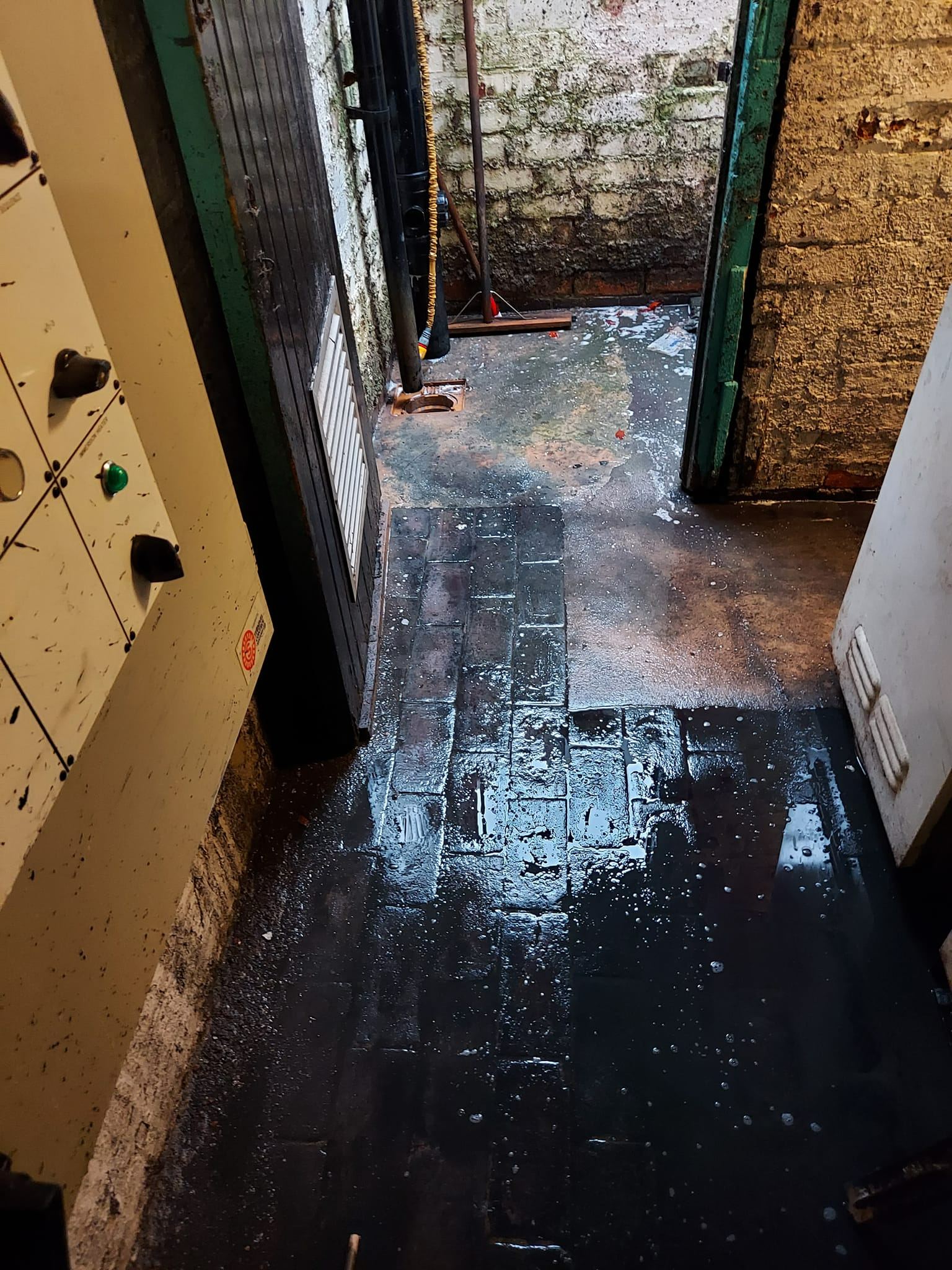 A dark, wet outdoor space with standing water and damp brickwork, suggesting a blocked drain or drainage problem.
