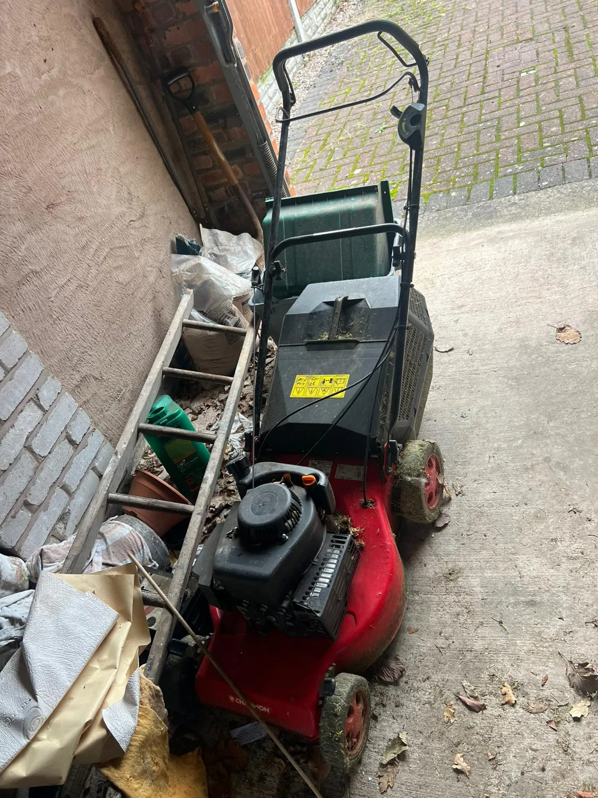 A messy garage or shed corner with an old lawnmower, ladder, and bags of rubbish for clearance.