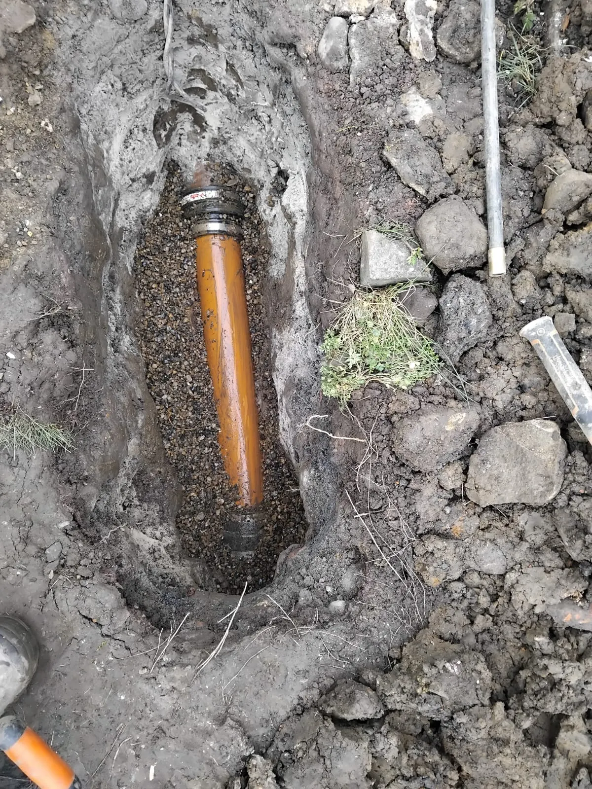 Orange drainage pipe installed in an excavated trench with gravel and soil, showing groundworks for a new drain.