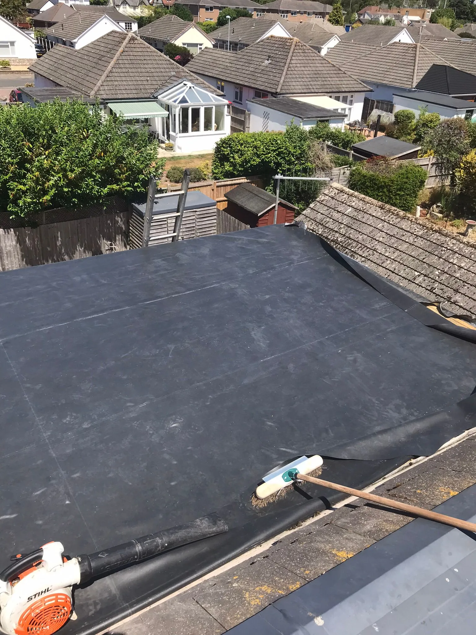 Flat roof installation in progress on a residential building, showing a dark roofing membrane, a Stihl leaf blower, and a broom, with other tiled house roofs in the background.