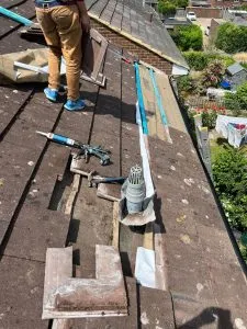 A roofer on a residential pitched roof, engaged in repair work, with removed tiles, tools, and a roof vent in focus.