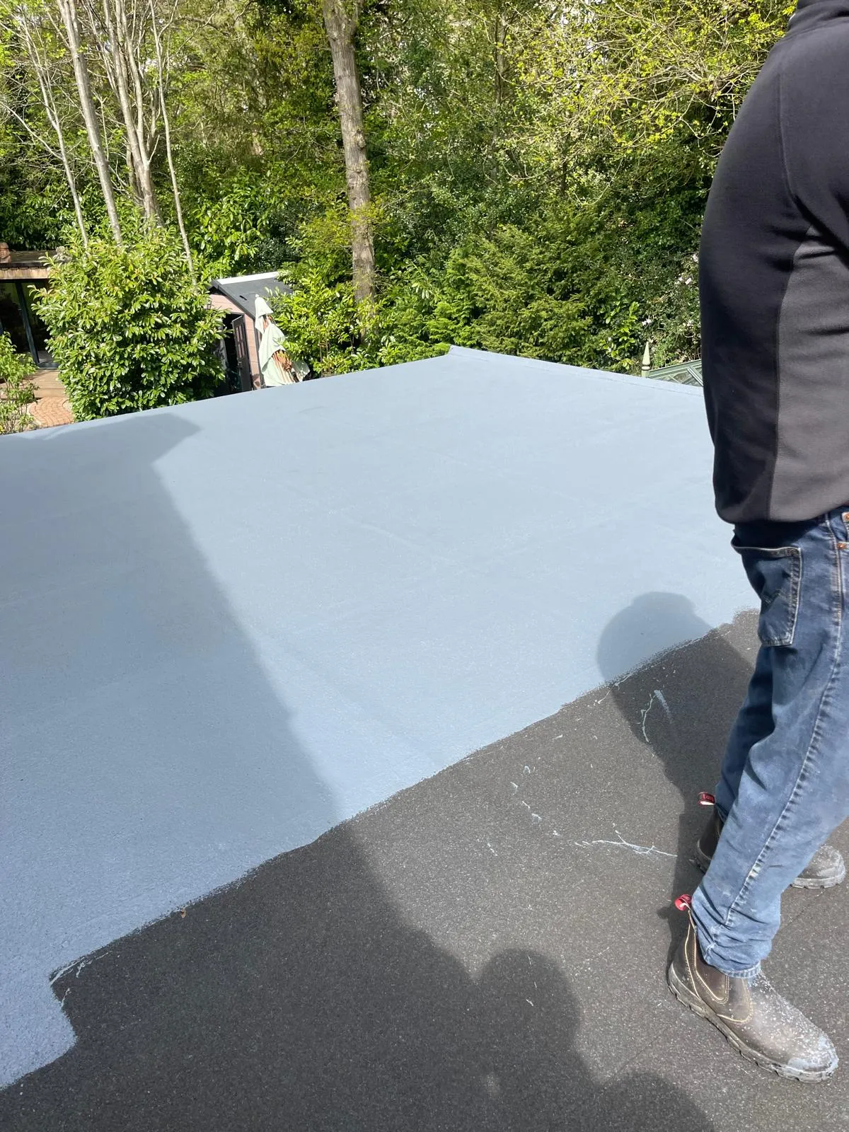 A section of a new light grey flat roof being installed, with a roofer's legs visible on the older roof surface.