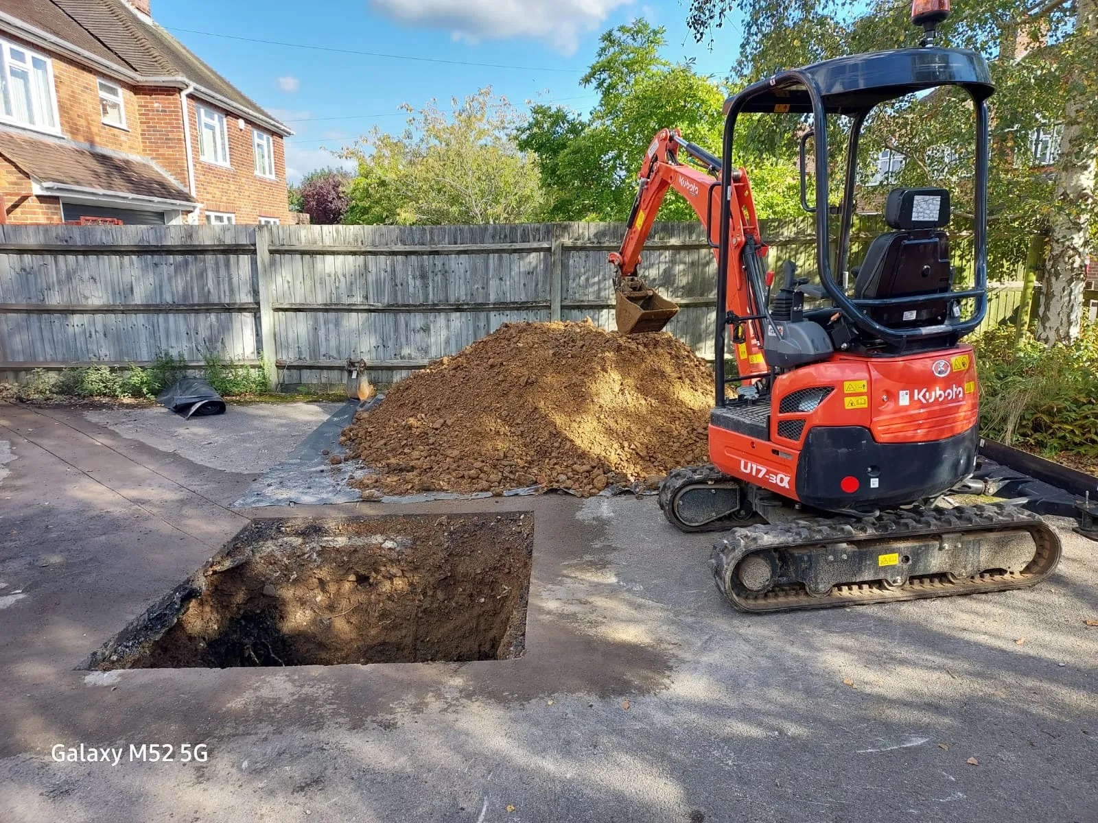 An orange Kubota mini excavator next to a large pit in a paved driveway, with a mound of earth beside it, indicating drainage repair or installation work.