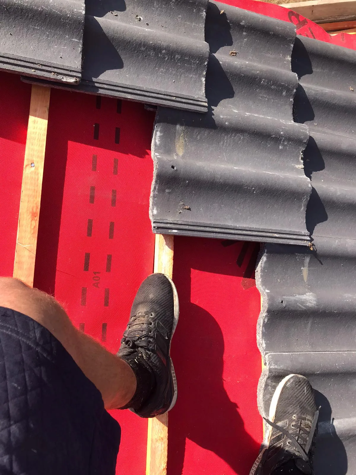 Roofer's feet on a roof undergoing installation, featuring new grey roof tiles, red underlayment, and timber battens.