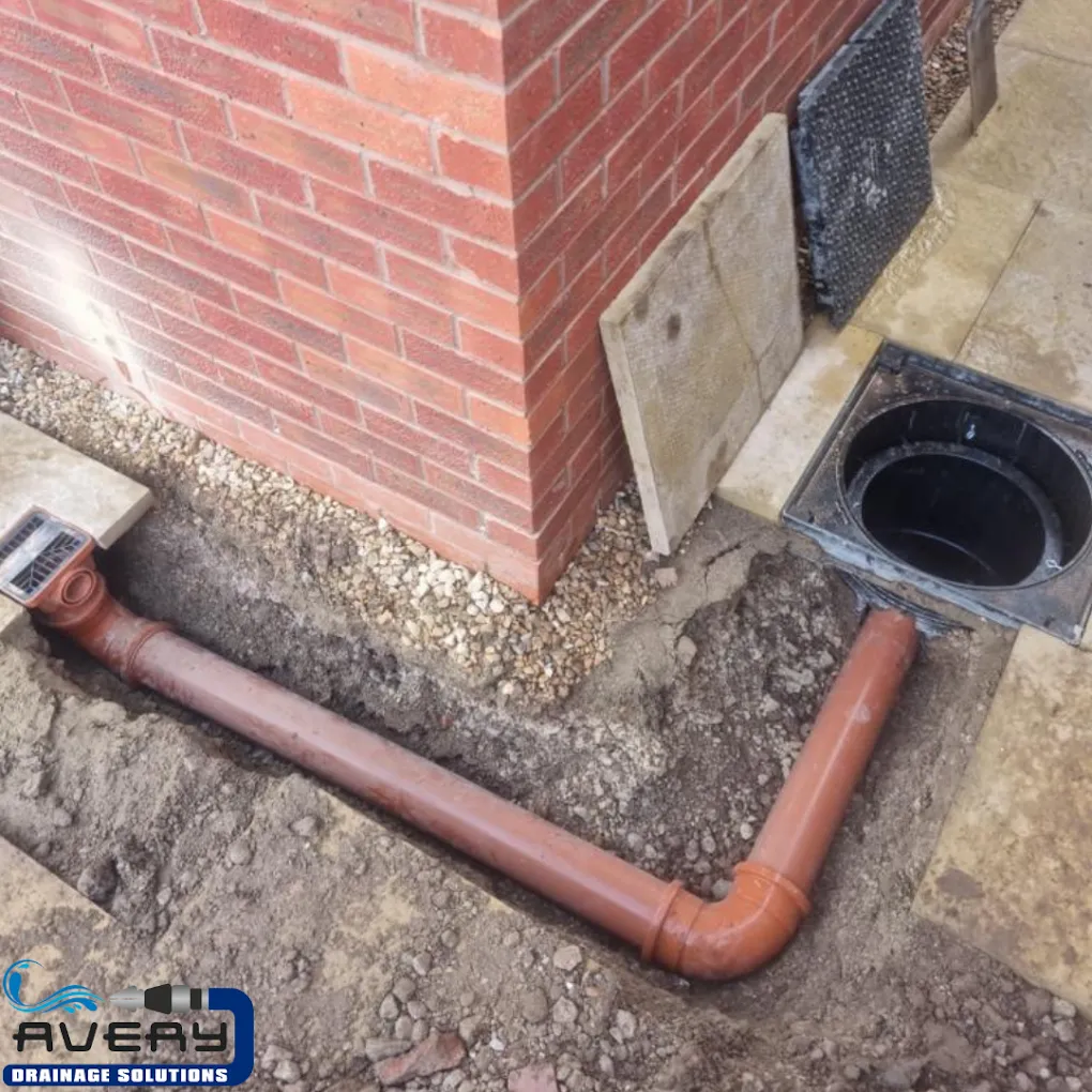 New drainage pipe and surface water drain being installed next to a brick building foundation.