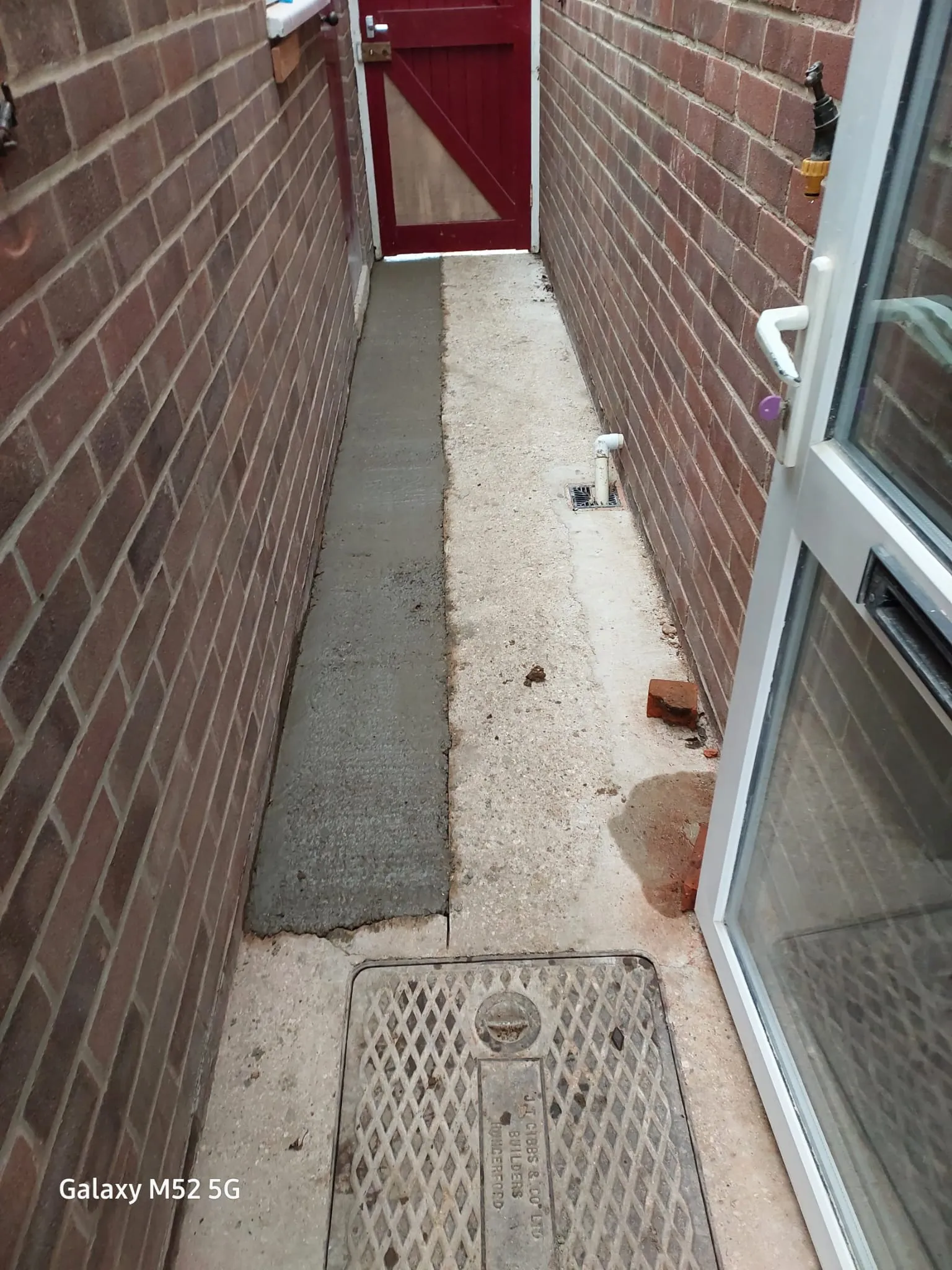 A residential side path with a drainage manhole cover, a new concrete strip, and a grate drain, indicating recent drainage system work.