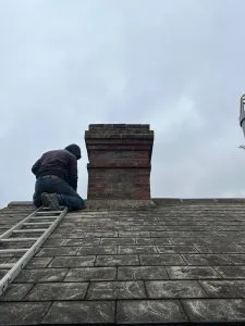 A roofer on a ladder inspecting or working on a brick chimney stack on a grey tiled roof.