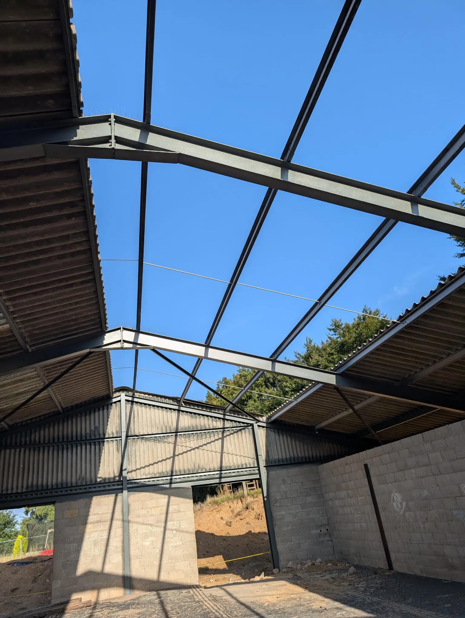 Interior of an industrial building with exposed steel roof frame and partial corrugated asbestos-style panels, against a bright blue sky.