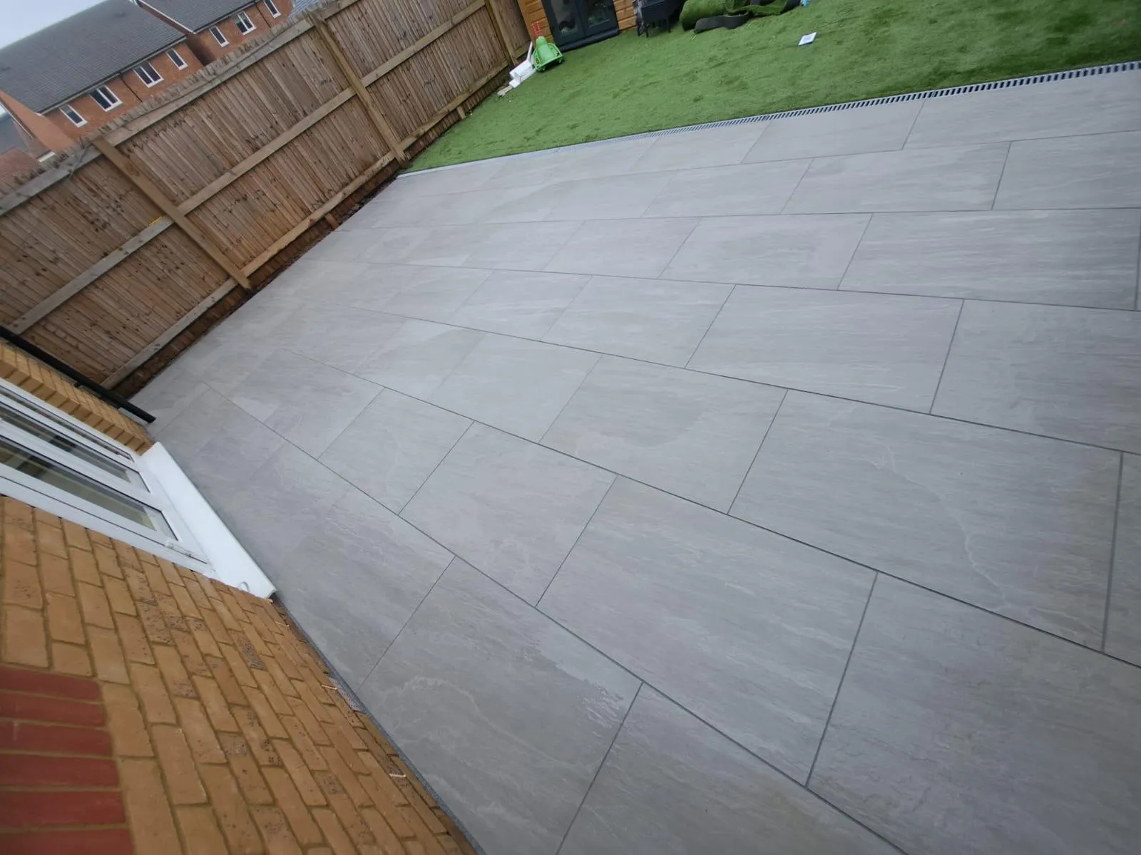 Modern light grey slab patio installation next to artificial grass and a wooden fence, showcasing a clean outdoor living space.