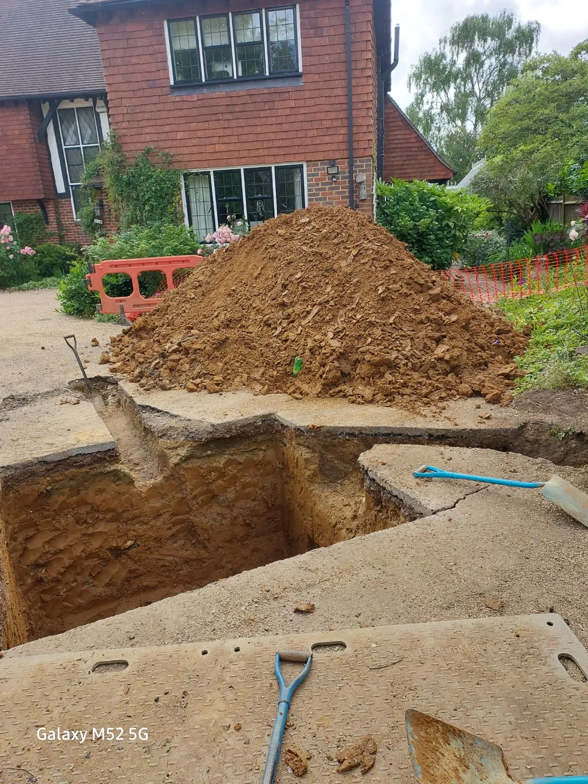 A construction site with a large trench dug for drainage repairs or installation, and a pile of excavated earth next to it, with a house in the background.