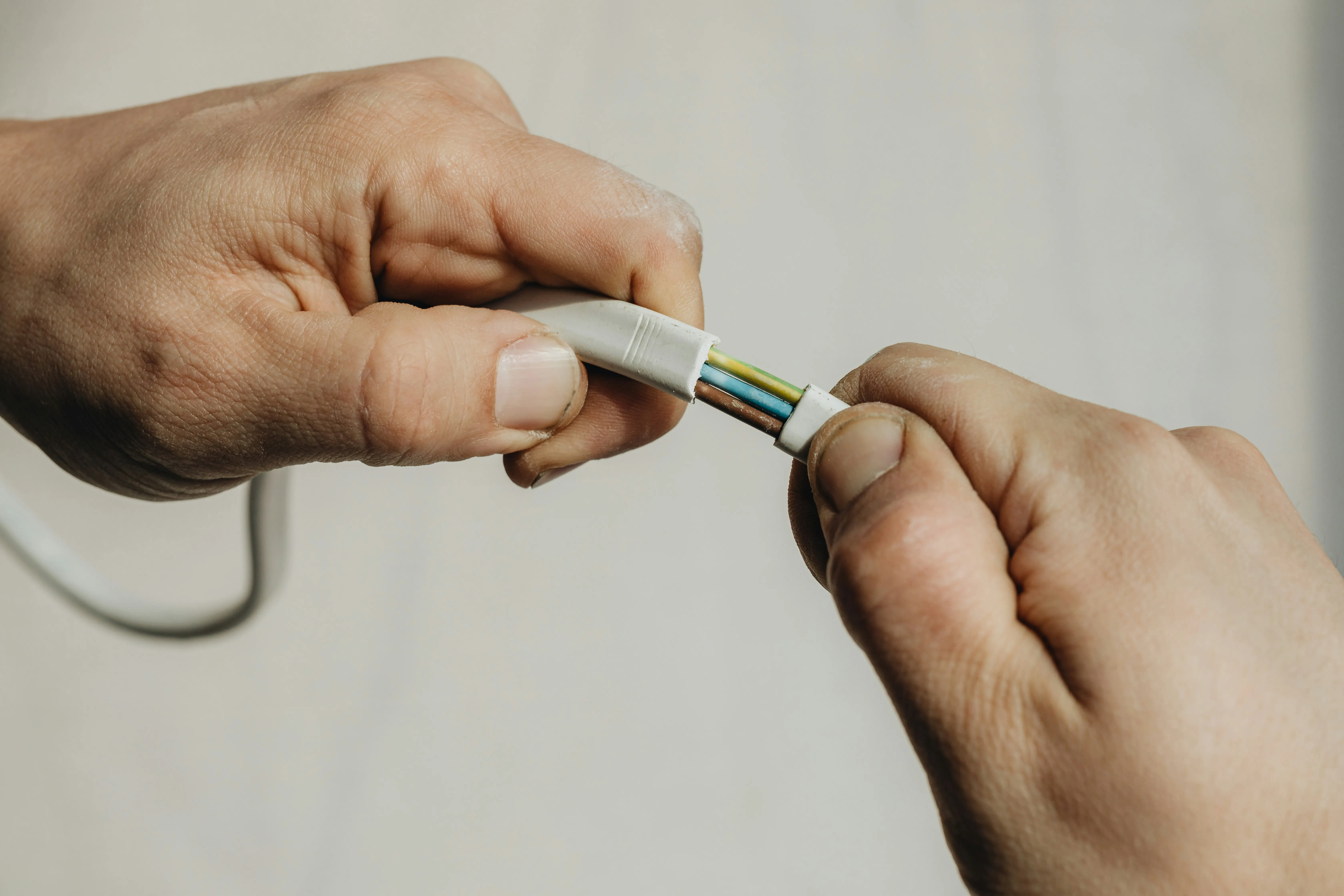 Close-up of an electrician stripping a white electrical cable to reveal the internal coloured wires (brown, blue, green/yellow).