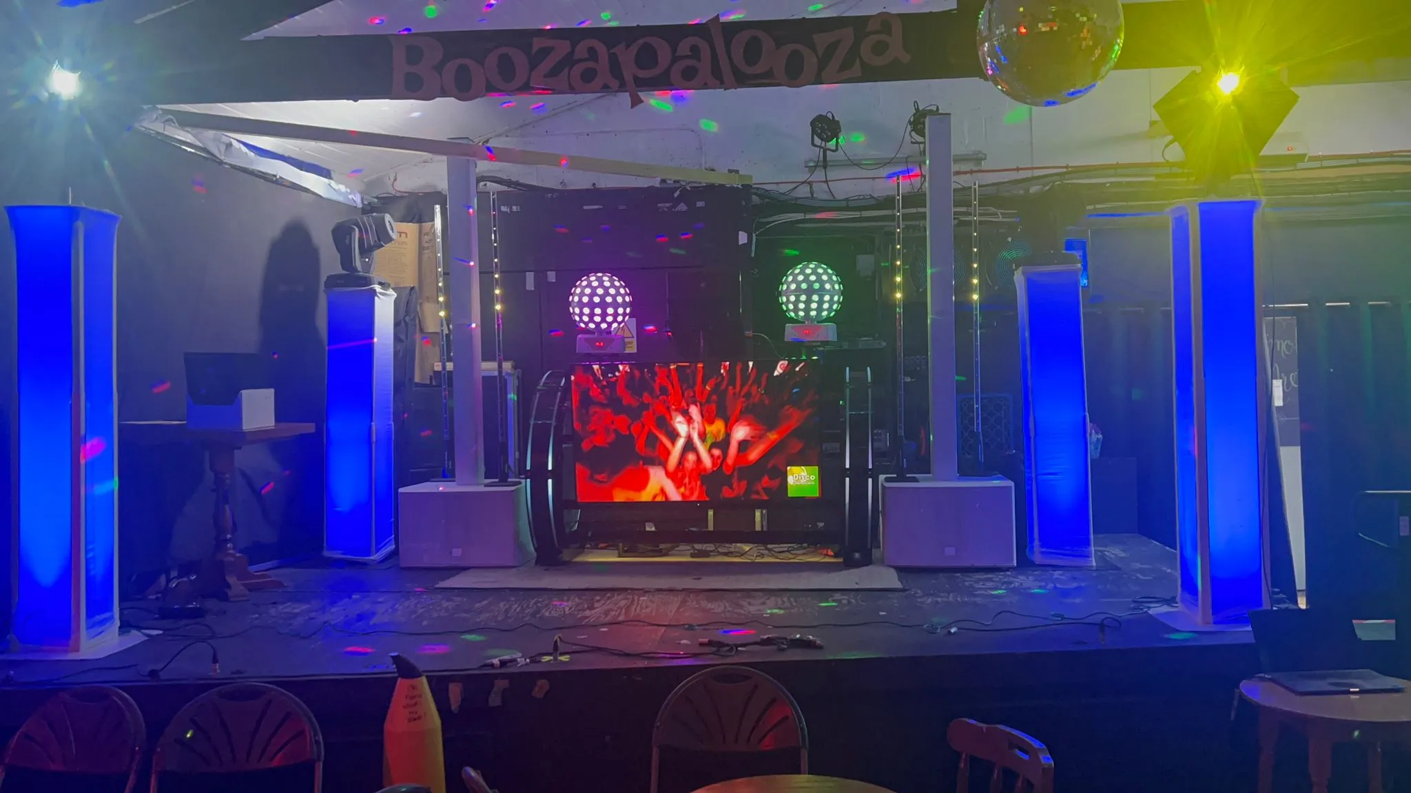Professional DJ entertainment setup on stage with a large video screen, blue LED lighting towers, and colourful disco effects, ready for a party.