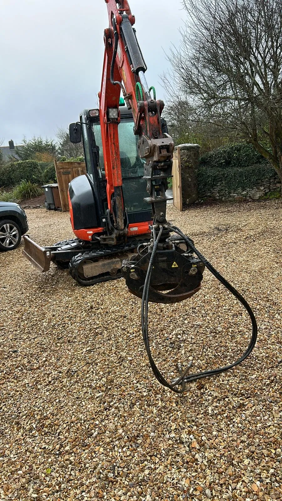 A mini excavator with a grab attachment on a gravel driveway, used for drainage installation and groundworks.