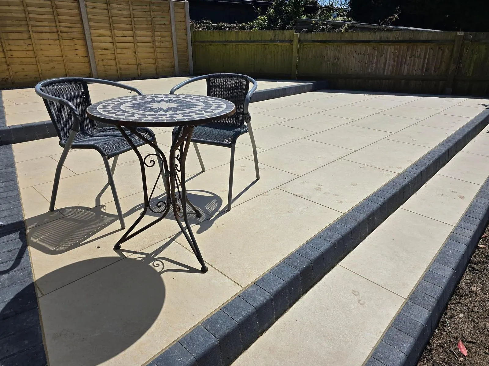 A modern patio with light paving slabs, dark grey block paving steps, and a mosaic outdoor table and chairs, set against a wooden fence.