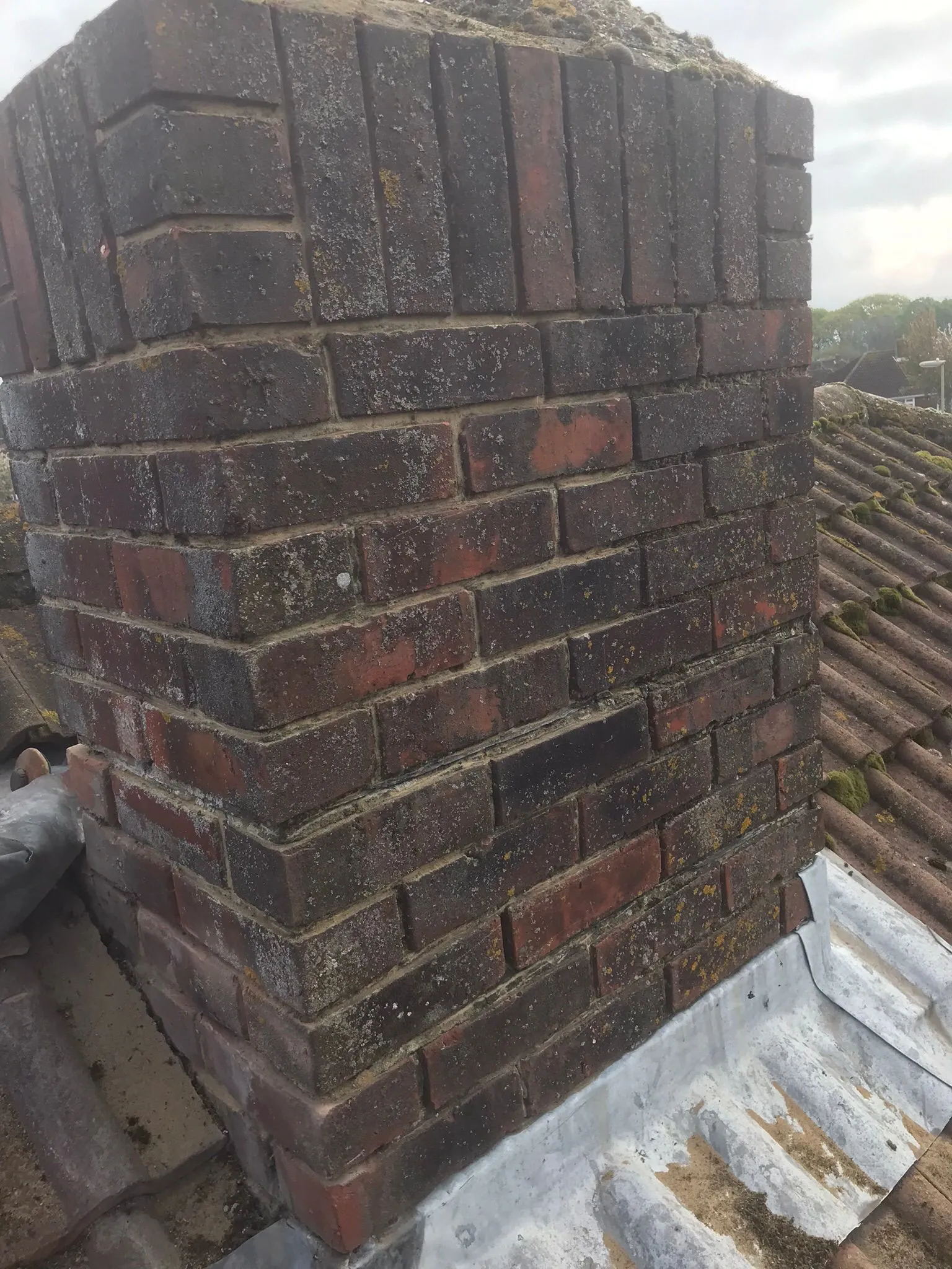 Weathered brick chimney on a tiled roof with moss and lead flashing, indicating a need for repair.