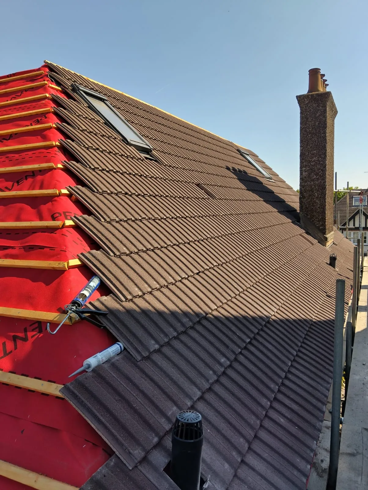Pitched roof installation in progress with brown tiles, red underlayment, skylights, and a chimney against a blue sky.