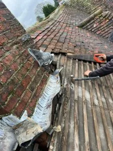 Roofer working on a damaged pitched roof, fixing missing tiles and old lead flashing next to a brick structure.