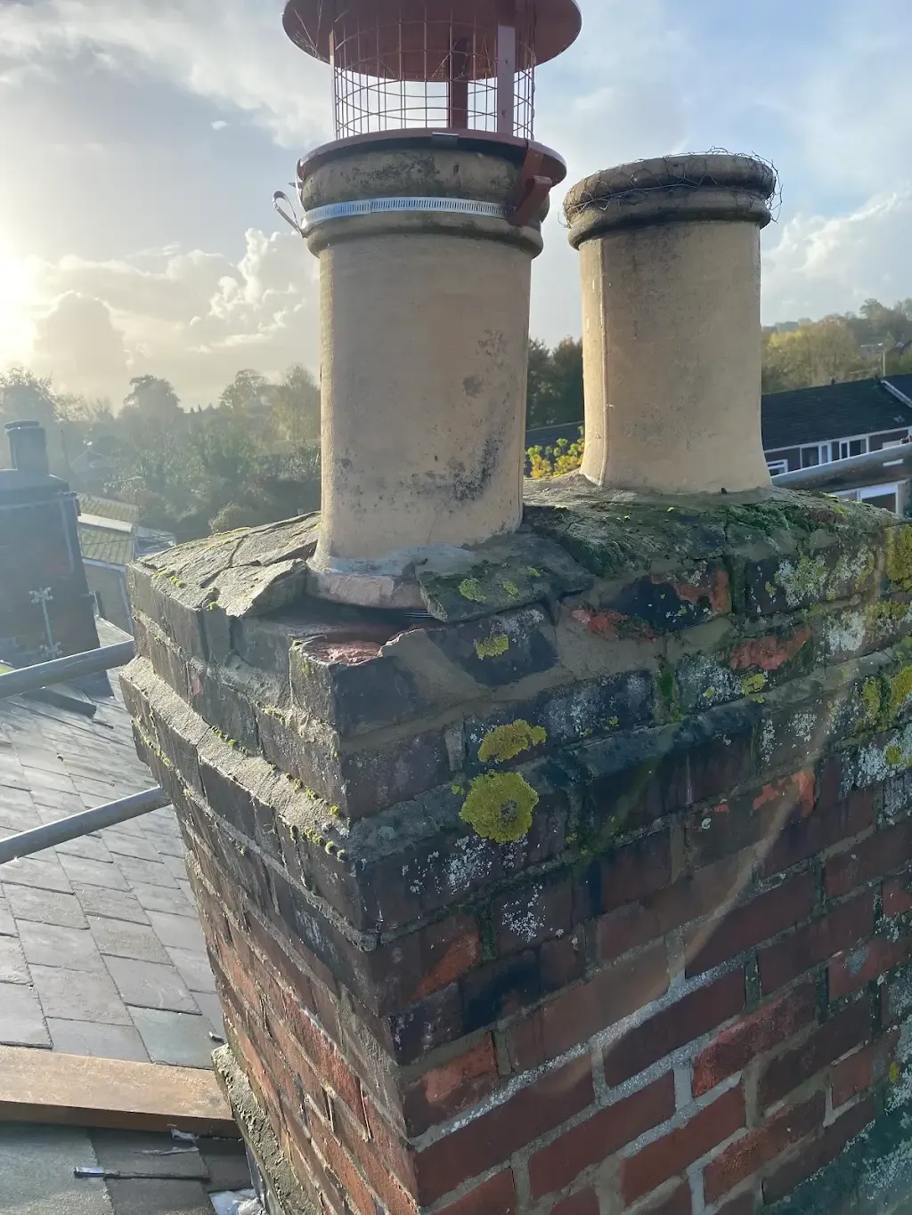 A damaged brick chimney with pots and a cowl on a slate tiled roof, showing cracks and moss.