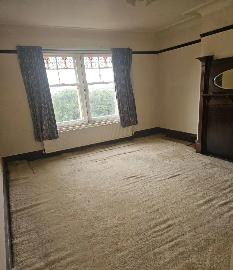 An empty room in an old house with a worn carpet, window, and fireplace, ready for house clearance or renovation.