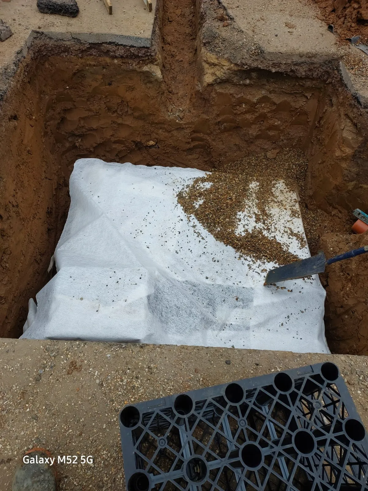 A newly installed soakaway crate, wrapped in white geotextile fabric and surrounded by gravel, within a deep trench excavation.