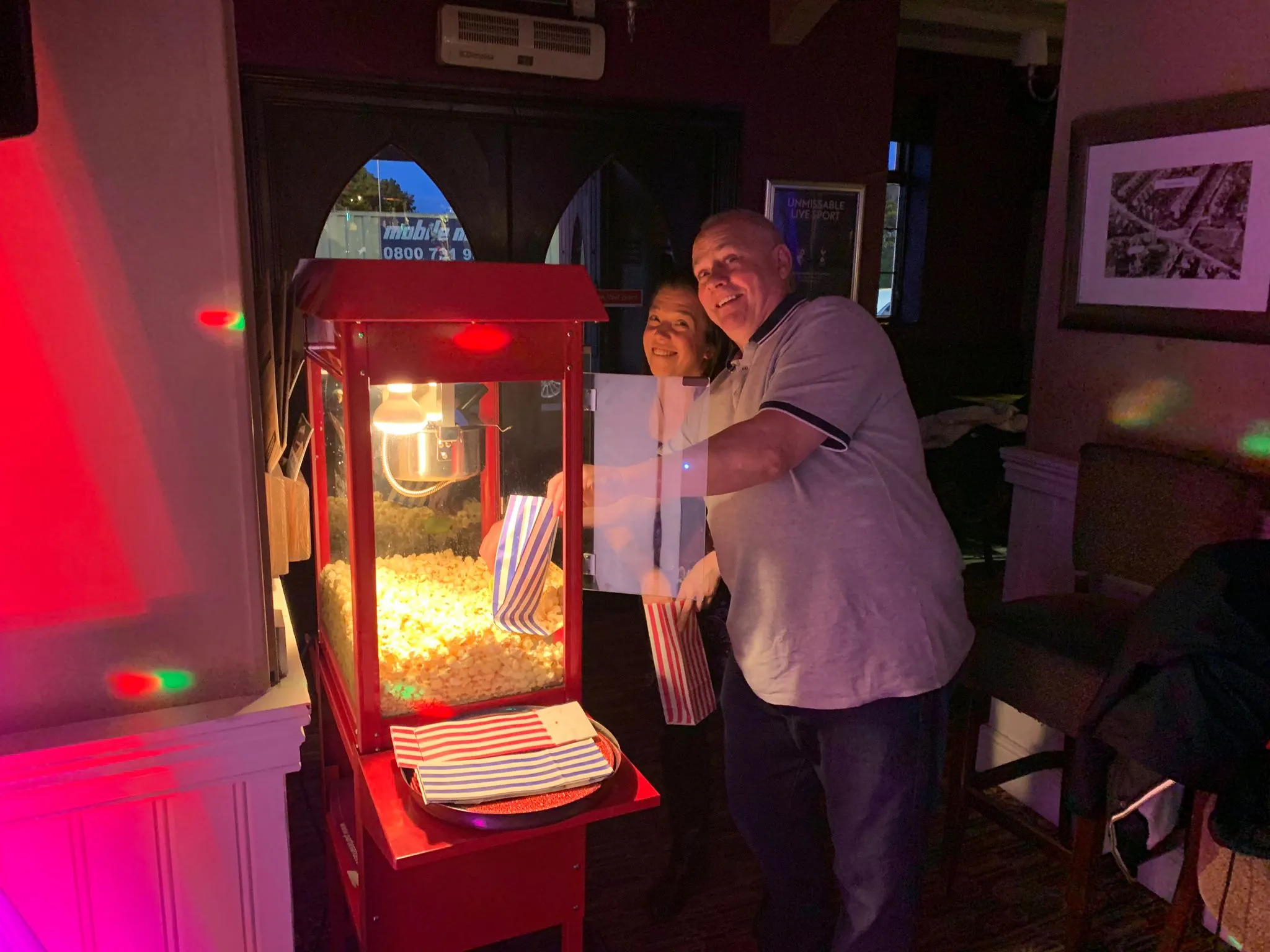 A man and woman smiling next to a vibrant red popcorn machine filled with popcorn and striped bags at an event.