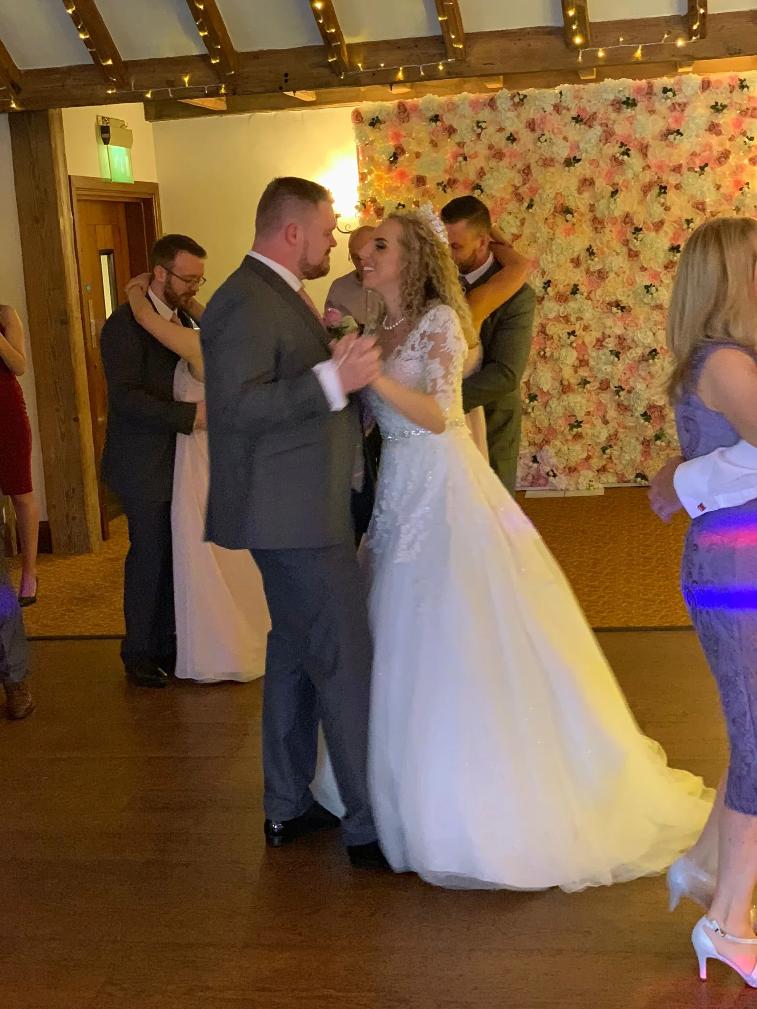 A smiling bride and groom share a dance at their wedding reception, surrounded by guests and the warm glow of event lighting, with a beautiful flower wall behind them.